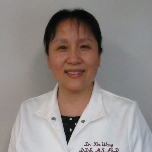 Smiling woman in a white lab coat standing against a light gray background; embroidered text on coat reads 'Dr. Xi Wan DDS MS PhD'